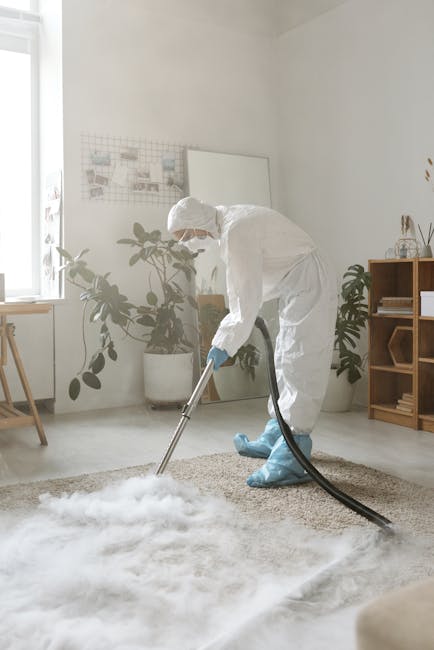 A professional cleaner dressed in full protective PPE, including a white coverall, face mask, goggles, and blue gloves, is using a steam cleaning machine to deep clean a beige carpet in a modern livin